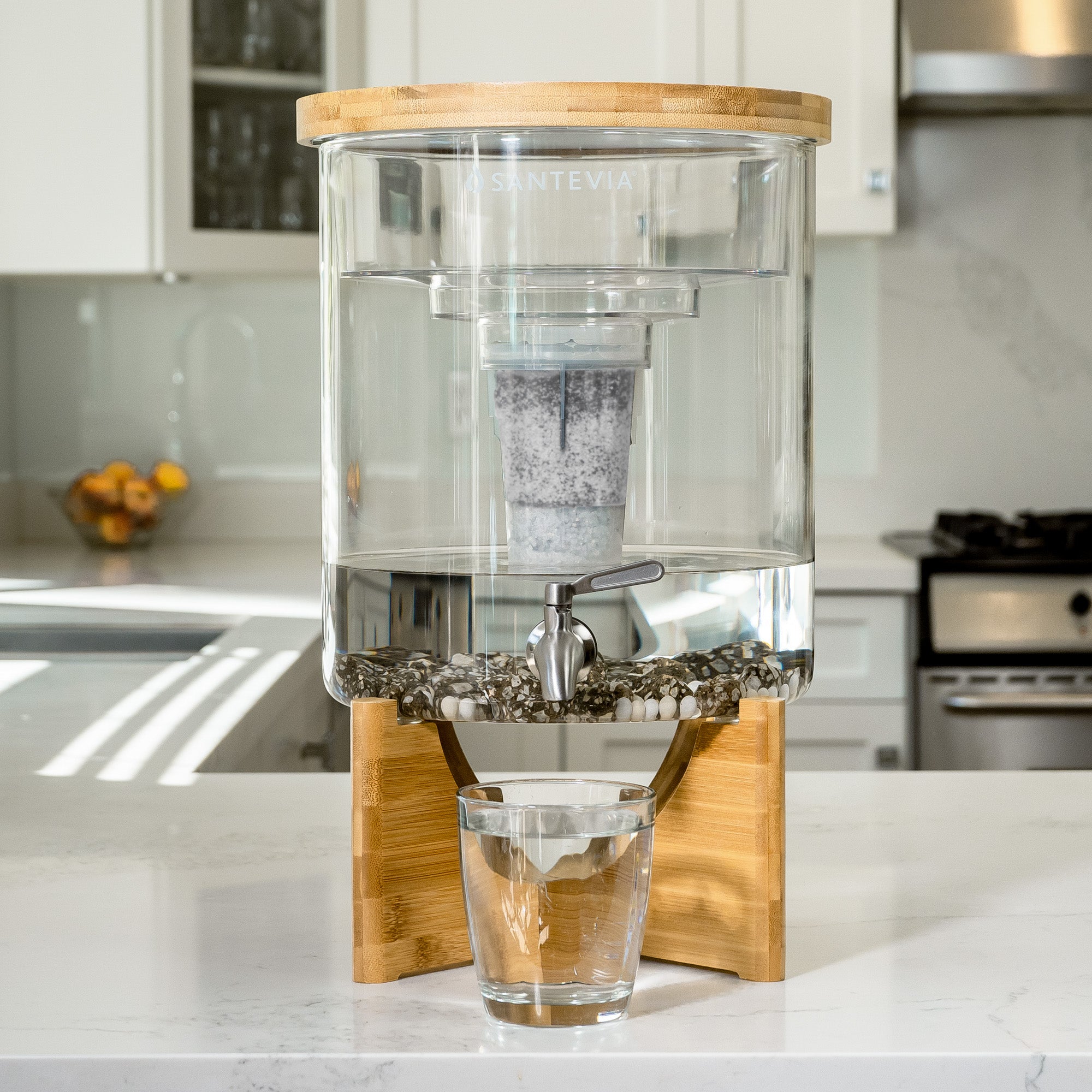 Clear glass water dispenser with wooden base and lid on a kitchen counter.
