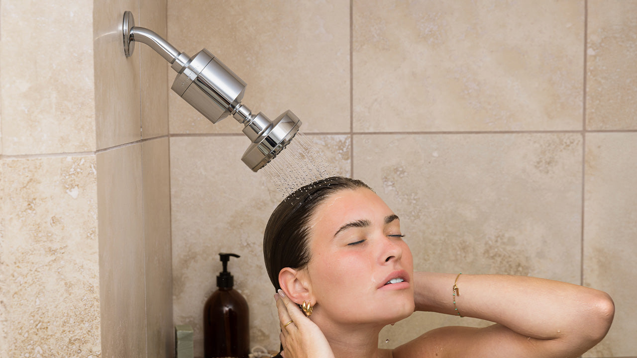 Woman showering with a shower head attached to the wall in a tiled bathroom.